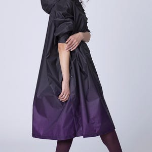 May include: A black hooded raincoat with a purple ombre design. The coat features a long, flowing silhouette and is paired with dark purple tights and black ankle boots. The coat has a subtle pattern.