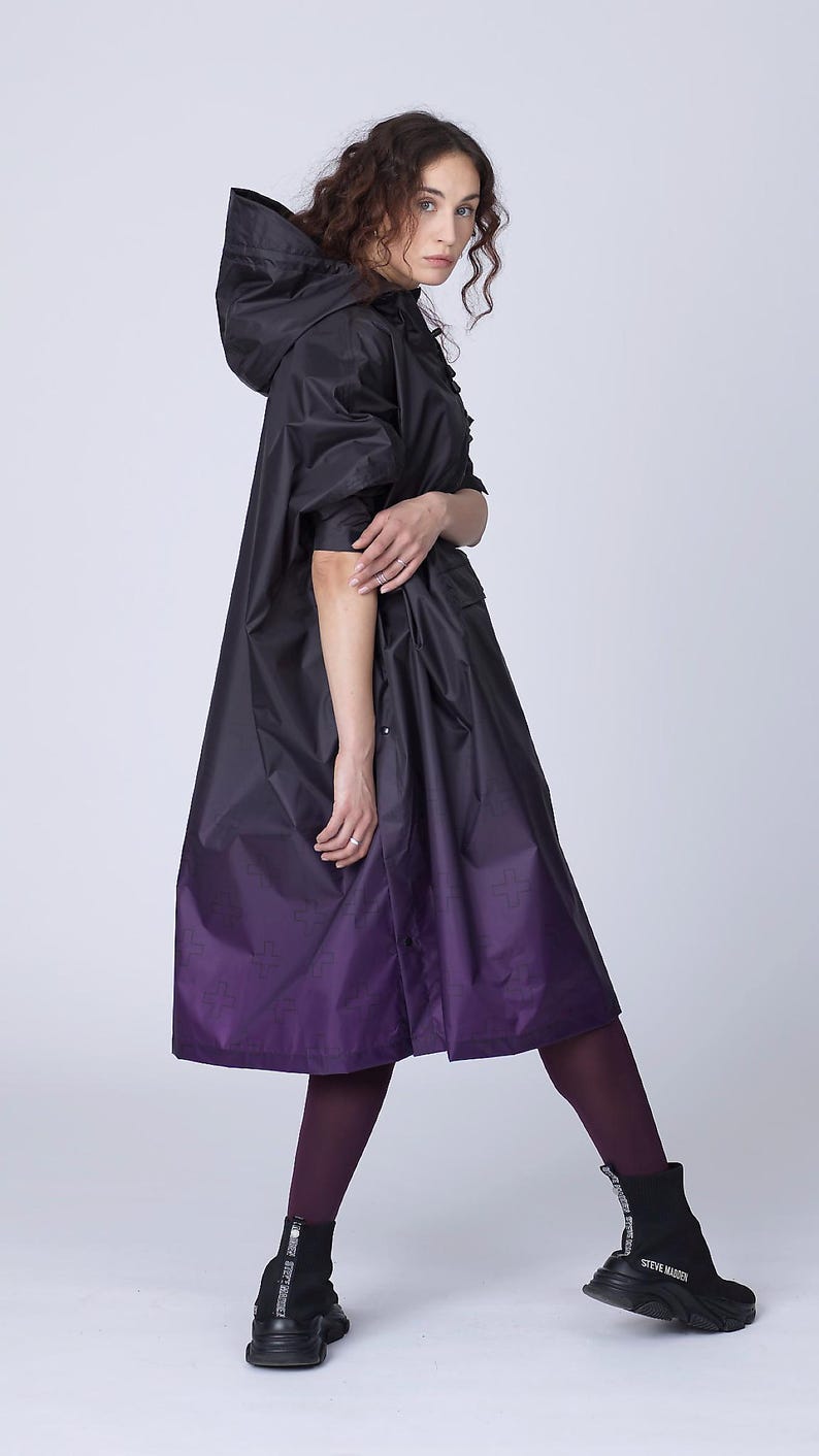 May include: A black hooded raincoat with a purple ombre design. The coat features a long, flowing silhouette and is paired with dark purple tights and black ankle boots. The coat has a subtle pattern.
