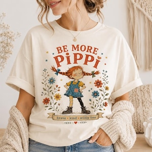 Könnte beinhalten: Cremefarbenes T-Shirt mit einer Illustration eines Mädchens mit roten Zöpfen, umgeben von Blumen. Der Text "BE MORE PIPPI" steht über der Illustration, darunter "brave • kind • wildly free". Die Ärmel sind hochgekrempelt.