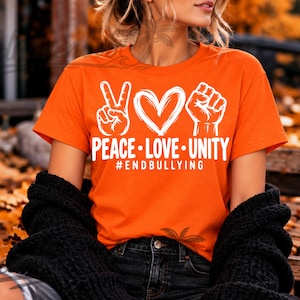 May include: A bright orange short-sleeved t-shirt featuring a white graphic print of a peace sign, a heart, and a raised fist. Below the icons, the text reads PEACE LOVE UNITY and the hashtag #ENDBULLYING in a bold, white sans-serif font.