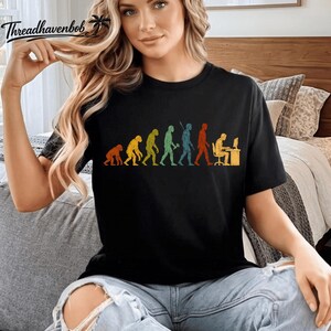 May include: A black short-sleeved t-shirt featuring a colourful graphic print that depicts the evolution of humanity, starting from a primate and progressing through various stages to a person sitting at a desk working on a computer.