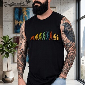 Puede incluir: Una camiseta sin mangas negra con un estampado gr&aacute;fico colorido de la evoluci&oacute;n de la humanidad, que progresa desde un primate hasta un humano sentado en un escritorio de computadora. El dise&ntilde;o utiliza una paleta de colores retro naranja, amarillo, verde y azul.