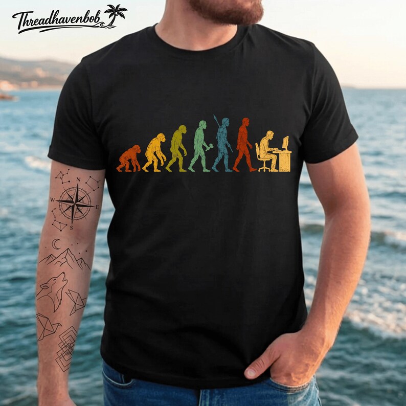 Puede incluir: Una camiseta negra de manga corta con un estampado gr&aacute;fico colorido de la evoluci&oacute;n humana, que comienza con un primate y termina con una persona sentada en un escritorio de computadora. El dise&ntilde;o utiliza una paleta de colores vintage.