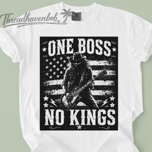May include: White t-shirt featuring a black and white graphic of a musician playing a guitar in front of an American flag design. The text "ONE BOSS NO KINGS" is printed above and below the graphic. The shirt has short sleeves.