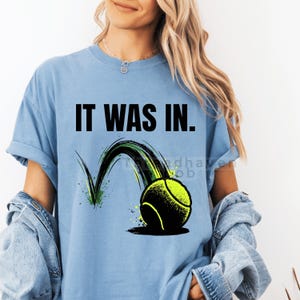 Puede incluir: Camiseta azul claro con el texto "IT WAS IN." y un gráfico de una pelota de tenis en movimiento. La pelota de tenis es amarilla y negra con un arco verde y negro detrás.