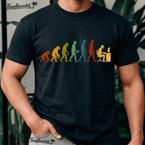 Puede incluir: Una camiseta negra de manga corta con un estampado gr&aacute;fico colorido de la evoluci&oacute;n humana. El dise&ntilde;o muestra la progresi&oacute;n desde un primate hasta un humano moderno sentado en un escritorio de computadora. El texto "Threadhavenbob" aparece arriba a la izquierda.