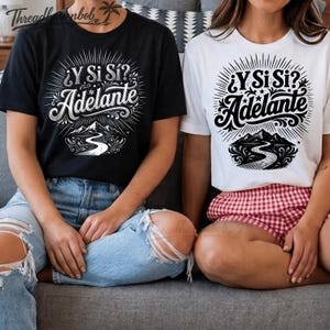 Peut inclure: Un t-shirt noir et un t-shirt blanc, chacun arborant une impression graphique avec le texte "¿Y Si Sí? Adelante" au-dessus d'une illustration stylisée d'une route sinueuse menant vers des montagnes, entourée de rayons de soleil et de volutes.