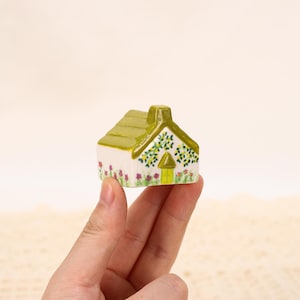 Può includere: Una piccola casa in ceramica con tetto e camino verdi, tenuta in mano. Le pareti bianche sono decorate con motivi floreali e di foglie, e una porta gialla. Un oggetto decorativo.