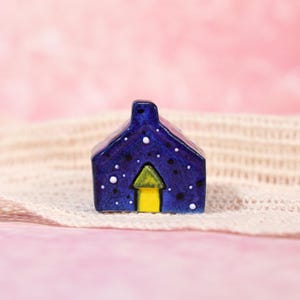 May include: A small, ceramic house figurine. The house is a deep blue with white and black speckles, resembling a starry night sky. The door and window are a bright yellow. The figurine is placed on a cream-colored, textured surface against a pink background.
