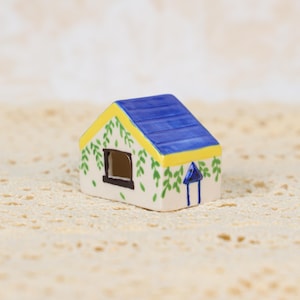 May include: A miniature ceramic house with a blue roof, yellow trim, and white walls adorned with green leaf patterns. The house has a small black window and a blue chimney. This decorative item is likely a collectible or a piece of home decor.