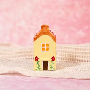 May include: A miniature yellow ceramic house with a brown roof, door, and orange window. Red flowers and green stems adorn the front. The house is set against a pink backdrop and a white, textured surface.