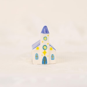 May include: A miniature ceramic church figurine. The white building has a blue roof and steeple, with yellow star and circle accents. Blue arched windows and a blue cross adorn the front door. A charming decorative piece.