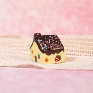 May include: A miniature ceramic house with a yellow exterior and a chocolate-colored roof. The roof is decorated with blue and red sprinkles. The house features strawberry and cookie designs, creating a whimsical, decorative piece.