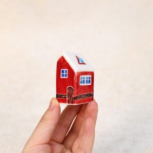 May include: A miniature red ceramic house with a white roof and blue window details. The house features a small door and two blue-framed windows. The house is held in a hand.