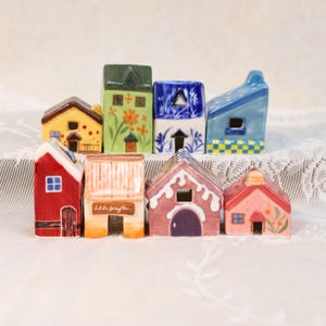 May include: A collection of colorful ceramic miniature houses. Each house features unique designs, including floral patterns, geometric shapes, and varying roof styles. The houses are painted in shades of red, yellow, green, blue, and pink.