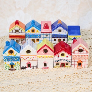 Puede incluir: Una colección de diez casas en miniatura de cerámica con diseños coloridos. Cada casa presenta un color de techo único, incluyendo rojo, azul y naranja, y detalles decorativos como ventanas y motivos florales. Las casas están dispuestas sobre una superficie de encaje.