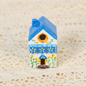 May include: A ceramic birdhouse with a blue roof and accents. The white body is decorated with yellow flowers and blue floral designs. A small, round opening is near the top, and a small door is at the bottom. The birdhouse is a decorative item.
