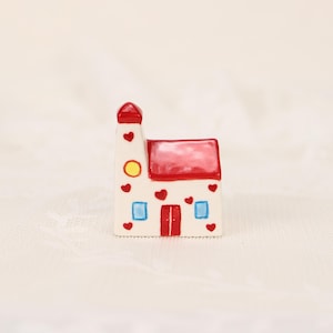 Può includere: Una piccola casa decorativa in ceramica con tetto e torre rossi. La casa bianca presenta cuori rossi e dettagli quadrati blu, un cerchio giallo e una porta rossa. Un affascinante oggetto decorativo.