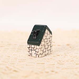 Könnte beinhalten: Eine Miniatur-Hausfigur mit grünem Dach und weiß gemusterten Wänden. Das kleine Haus hat einen schwarzen Schornstein und eine weiße Tür. Die Figur steht vor einem cremefarbenen, strukturierten Hintergrund.