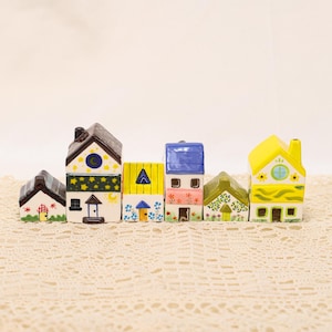 May include: A collection of colorful ceramic houses with varying designs. Each house features unique details, including star and moon accents, floral patterns, and different roof colors. The houses are arranged on a textured, off-white surface.