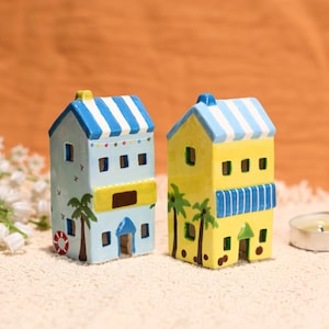 May include: Two ceramic miniature houses, one light blue and one yellow, with blue and white striped roofs. The light blue house has a red and white life preserver, and a palm tree painted on the side. The yellow house has palm trees painted on the side.