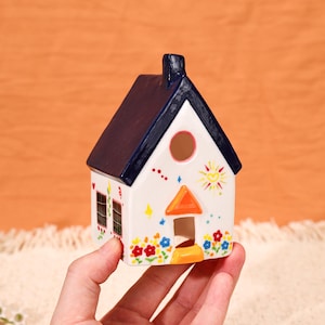 May include: A white ceramic house with a dark blue roof, chimney, and window frames. The house is decorated with colorful flowers, a sun, and stars. The house has a round window and an orange door.