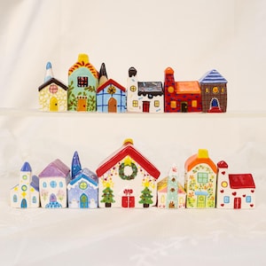 Op de afbeelding: Een verzameling kleurrijke keramische huizen, waaronder kerken en huisjes. De huizen hebben heldere kleuren, gedetailleerde ramen en decoratieve elementen. De huizen zijn gerangschikt op een wit oppervlak.