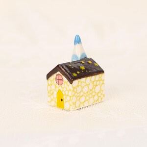 May include: A miniature ceramic house with a yellow and white dotted exterior, a brown roof with yellow star accents, and a blue and white chimney. The house has a yellow door and a pink window.