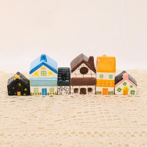 May include: A row of miniature ceramic houses in various colors. The houses have different roof styles and decorative details, including stars, windows, and doors. The color palette includes blue, yellow, brown, and pink.