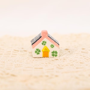 Könnte beinhalten: Ein Miniatur-Keramikhaus mit rosa Dach, Schornstein und Türrahmen. Das Dach ist schwarz, und das Haus ist weiß mit Kleeblatt-Designs. Die Tür ist gelb und orange.