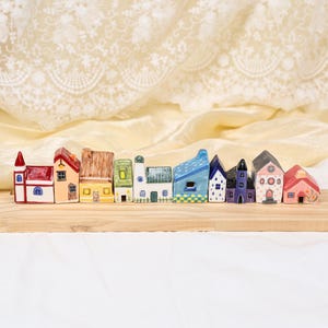 Puede incluir: Una colección de casas de cerámica en miniatura en varios colores, incluyendo rojo, amarillo, azul y verde. Cada casa tiene detalles arquitectónicos únicos, como chimeneas, ventanas y puertas. Las casas están dispuestas sobre una superficie de madera.