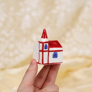 Könnte beinhalten: Eine Miniaturkirche aus Keramik mit rotem Dach und Turm sowie weißen Wänden. Die Kirche hat rote Verzierungen und blaue Bogenfenster. Das kleine Gebäude wird in der Hand einer Person gehalten.
