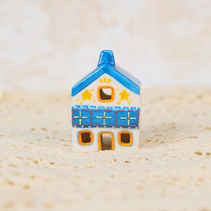 May include: A small, ceramic house figurine with a white base, blue roof, and blue accents. The house features yellow star and square window details. The roof has a chimney and the windows are outlined in orange.