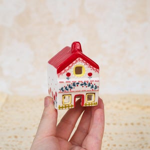 May include: A small, decorative ceramic house with a red roof and white walls. The house is adorned with floral patterns in red, green, and blue. It features yellow windows and a red door, creating a charming, miniature cottage aesthetic.