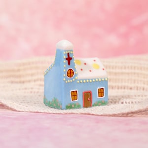 May include: A miniature blue church figurine with a white steeple and a red cross. The roof is white with yellow and red polka dots. The church has two orange windows and a brown door. The base of the church has small green and pink floral accents.