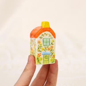 May include: A small ceramic house with a yellow chimney, orange roof, and a green door and window. The house is decorated with yellow and orange flowers and green vines. The house is held in a hand.