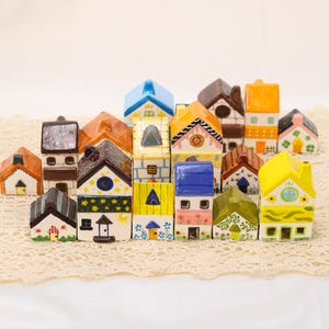 May include: A collection of miniature ceramic houses in various colors and designs. The houses have different roof colors, including blue, orange, and brown. Some houses feature decorative details like stars, moons, and floral patterns. The houses are arranged on a lace surface.
