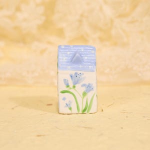 May include: A small, ceramic house figurine with a light blue roof and white walls. The roof has a triangle peak and is dotted with white spots. The walls are decorated with blue floral designs and green stems.