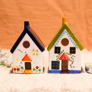Puede incluir: Dos portavelas de cerámica con forma de casa. Uno tiene un tejado azul, una puerta naranja y un diseño de sol. El otro tiene un tejado verde, una puerta roja y detalles florales. Ambos tienen aberturas circulares y ventanas cuadradas.