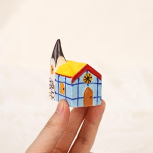 May include: A small, hand-painted wooden church figurine. The church has a blue body with a yellow roof and a black steeple. The details include a red trim, an orange door, and a star design.