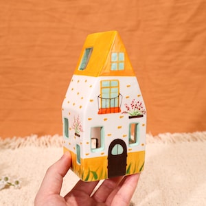 May include: A ceramic house figurine with a yellow roof and accents. The white body is decorated with orange polka dots, windows, and a balcony with flowers. The door is brown, and the base has green grass details.