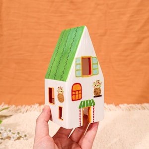 May include: A white ceramic house with a green roof, windows, and a door. The house has a decorative design with flower baskets and a red and white striped awning over the door. The house is held in a hand.