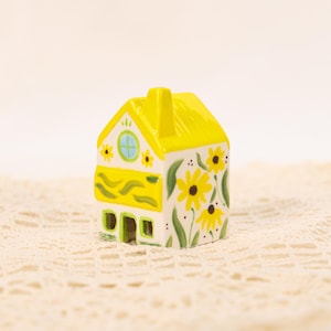 May include: A miniature ceramic house with a yellow roof and floral detailing. The house is painted white with yellow sunflowers and green leaves. The house has a small, round window and green trim.