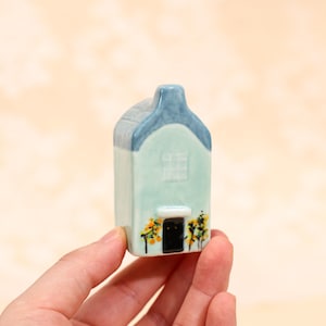 May include: A miniature ceramic house with a light blue facade and a dark blue roof. The house features a small black door, a white window, and yellow and green floral accents. The house is held in a hand.