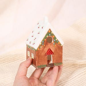 May include: A ceramic gingerbread house with a white roof decorated with red cherries and green garland. The house has a brown body, small windows, and a red door. The house is approximately 4 inches tall.