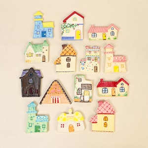Puede incluir: Una colección de catorce pequeños adornos de casa de cerámica, planos y pintados a mano, en varios estilos arquitectónicos y colores, incluyendo azul, amarillo, rosa, verde y marrón, con ventanas, puertas, techos y patrones pintados.