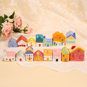 May include: A collection of colorful ceramic miniature houses with various roof designs and painted details. The houses are arranged in two rows, showcasing a variety of colors including pink, blue, orange, and green. The background features a floral arrangement and lace.
