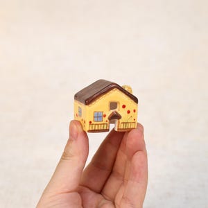 May include: A miniature ceramic house with a brown roof, yellow walls, and red dot accents. The house features a small chimney, blue window, and a decorative porch. The house is held in a hand.