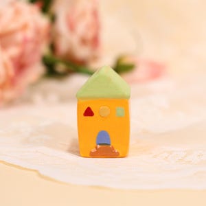 May include: A miniature ceramic house with a bright yellow body, a green roof, and a blue door. The house features a red triangle, a yellow circle, and a green square as decorative elements. The house is a charming decorative item.