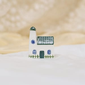 Peut inclure: Une petite maison en céramique blanche avec un toit vert et des détails bleus. La maison a une petite tour, une porte d'entrée et deux fenêtres. Une petite clôture longe la base. Objet décoratif.
